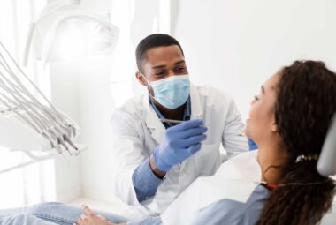 A hygienist and a patient in a bright operatory. The African American hygienist has paused his efforts while his patient answers a question. Behind his mask, you can tell the hygienist is smiling at his patient's response.