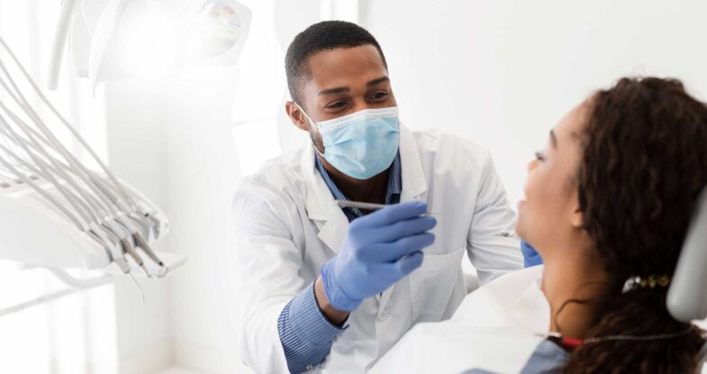 A hygienist and a patient in a bright operatory. The African American hygienist has paused his efforts while his patient answers a question. Behind his mask, you can tell the hygienist is smiling at his patient's response.